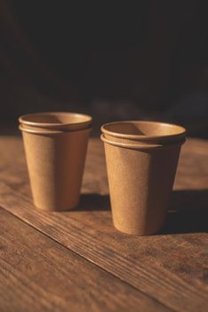 Disposable dark brown kraft paper cups stand on wooden table against brown background