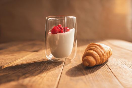 double glass with yogurt and berries and croissant on a wooden table where the bright sun is shining