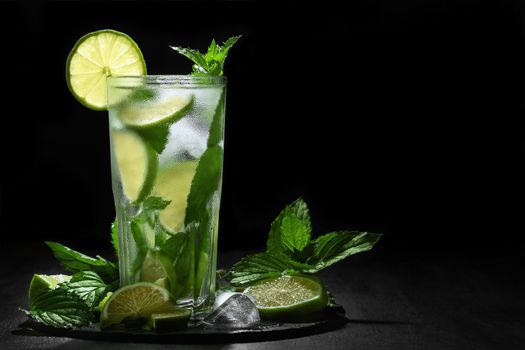 Fresh homemade mojito cocktail in a tall glass with lime, mint and ice on a black background, copy space.