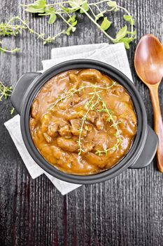 Beef goulash in tomato sauce with sprigs of thyme in a pan on a napkin, spoon, parsley on black wooden board background from above