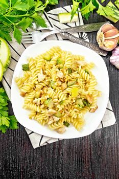Fusilli pasta with chicken breast, zucchini, cream and pine nuts in a plate on napkin, garlic, fork and parsley on a dark wooden background from above
