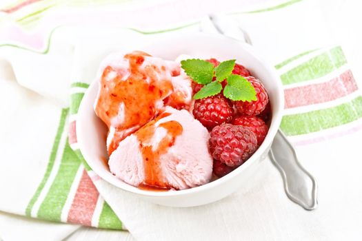 Ice cream crimson with raspberry berries, syrup and mint in white bowl, a spoon on towel against light wooden board