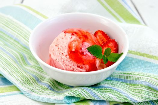 Strawberry ice cream in a white bowl with strawberries and strawberry syrup on a striped linen napkin on a wooden boards background