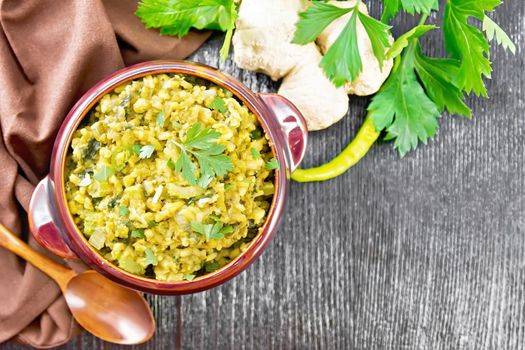 Indian national dish of kichari of mung bean, rice, celery, spinach, hot pepper and spices in a bowl on a napkin, ginger and spoon on dark wooden board background from above