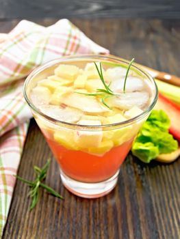 Lemonade with rhubarb and rosemary in a glass, stems and a leaf of a vegetable, napkin on a background of a dark wooden board