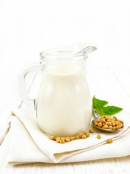 Soy milk in a jug, green leaf, soybeans in a spoon on a napkin against the background of a light wooden board

