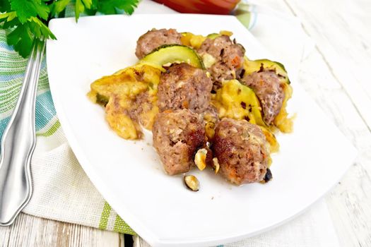 Meatballs baked with zucchini, cheese and nuts in a dish on a towel, parsley and fork on a light wooden board background