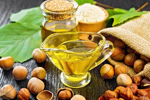 Hazelnut oil in a glass jar and gravy boat, flour in a bowl, nuts in a bag, spoon and on the table, filbert sprigs with green leaves on wooden board background