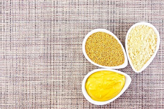 Mustard sauce, seeds and mustard powder in three saucepans on a background of brown woven napkin on top