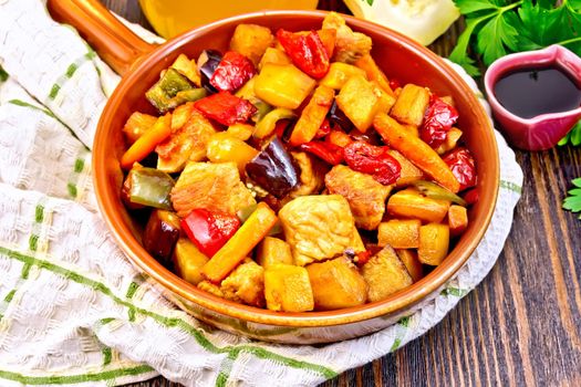 Roast of meat, zucchini, eggplant, carrot and sweet pepper with honey, soy sauce and red wine in a ceramic pan on a napkin against the background of wooden boards