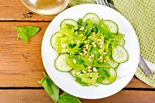 Salad from spinach, fresh cucumbers, rukkola salad, cedar nuts and spring onions, seasoned with vegetable oil on a plate, napkin and fork on a wooden board background from above