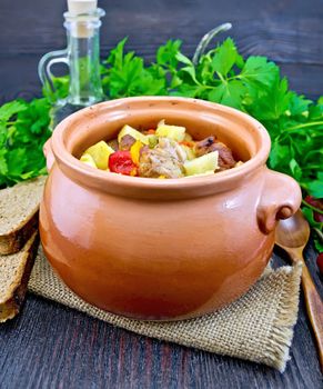 Roast with chicken, potatoes, squash and sweet peppers in a clay pot on a napkin of burlap, spoon, parsley on a dark wooden board