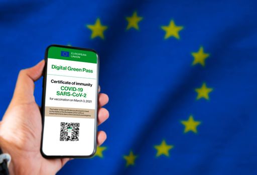 The digital green pass of the european union with the QR code on the screen of a mobile held by a hand with a blurred EU flag in the background. Immunity from Covid