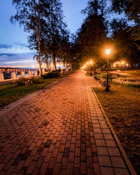 A night park lit by lanterns with a stone pavement, trees, fallen leaves and benches in early autumn. Cityscape.