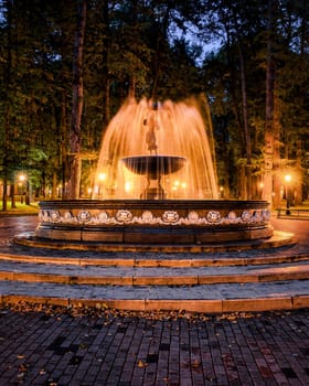 A fountain with blurred streams of water in a night park illuminated by lanterns with a stone pavement, trees and benches. Cityscape.