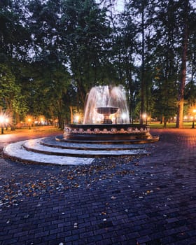 A fountain with blurred streams of water in a night park illuminated by lanterns with a stone pavement, trees and benches. Cityscape.