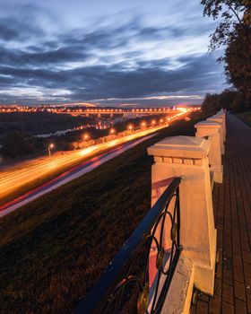 Moving car with blur light through city at night. Bridge over the river and the road. A view from the park from a height with a fence in the foreground. Cityscape.