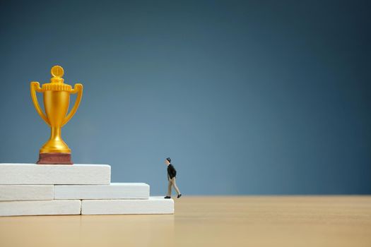 Miniature business concept - businessman walking on white staircase ladder to reach golden trophy. Image photo