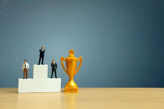 Miniature business concept - businessman standing in front of white winner podium with golden trophy. Image photo