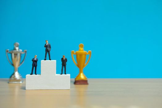 Miniature business concept - three businessman standing on winner podium with golden and silver trophy. Image photo