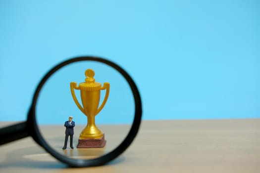 Miniature business concept - businessman standing in front of golden trophy and magnifier glass. Image photo
