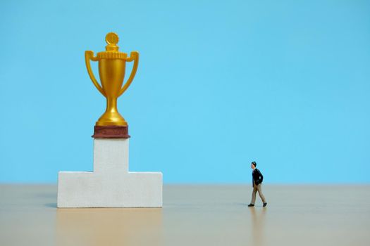 Miniature business concept - businessman walking toward golden trophy above white podium. Image photo