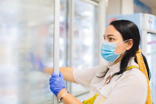 Woman wearing a protective face mask and disposable gloves browsing the frozen food section while shopping at a supermarket, selective focus