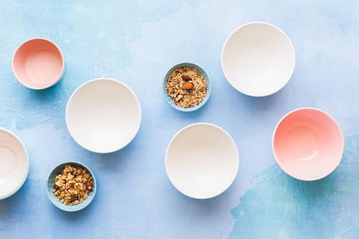 Granola crumbs in a bowls among empty ceramic multicolored  bowls on rustic blue background. Top view, blank space