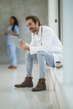 Shot of a mature doctor using digital tablet while sitting near a window in a hospital hallway during the Covid-19 pandemic.