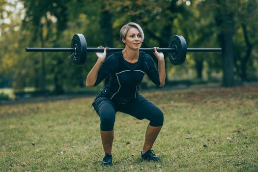 A fit mature woman is doing squat exercises, dressed in a black suit with an EMS electronic simulator to stimulate her muscles.