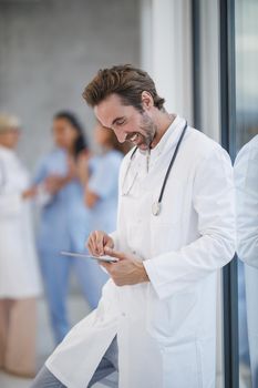 Shot of a smiling doctor standing near a window and using digital tablet while having quick break in a hospital hallway.