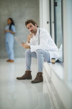 Shot of a mature doctor using digital tablet while sitting near a window in a hospital hallway during the Covid-19 pandemic.