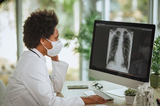 Shot of a worried African female doctor with protective N95 mask sitting alone in her consulting room and looking X-ray  of lungs on computer during COVID-19 pandemic.