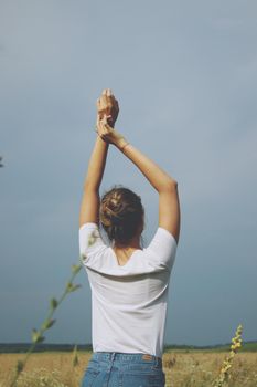 A blonde girl in white t-shirt and jeans standing in the field hands up with her back to us