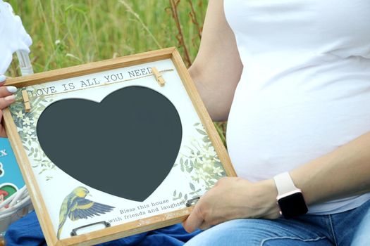 Pregnant woman in white t-shirt holding a little blank heart - shaped board in the park