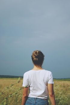 A blonde girl in white t-shirt and jeans standing in the field with her back to us