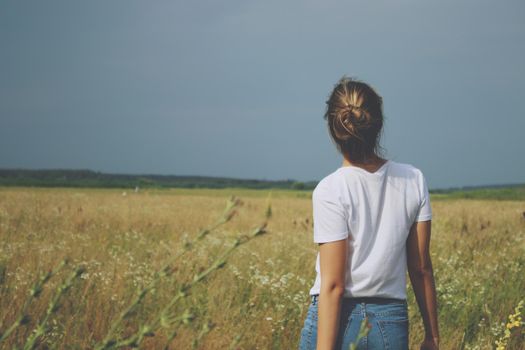 A blonde girl in white t-shirt and jeans standing in the field with her back to us