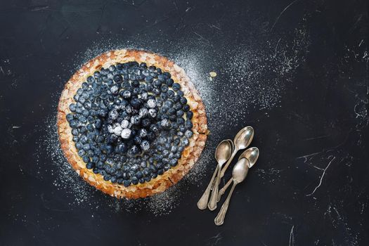 Delicious homemade blueberry almond tart with fresh blueberries and bunch of teaspoons beside. Top  view, blank space, dark toned image