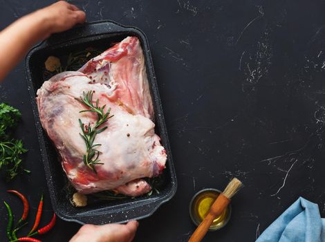 Woman preparing a meat roast with herbs and garlic. Ready to roast shoulder of lamb seasoned with herbs in a roasting tin. Top view, blank space
