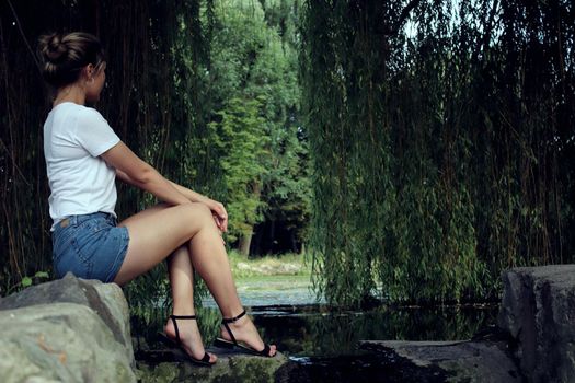 A blonde girl in white t-shirt and jeans sitting on a ground in front of a tree near the lake
