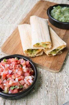 Tamales fresh out of the steamer on a wooden cutting board with pico de gallo and salsa verde toppings.