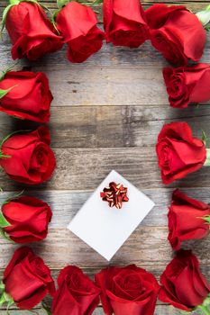 Frame of red roses and white gift box on wooden surface.