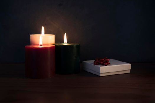 Three candles on a wooden surface with white gift box topped with red ribbon.
