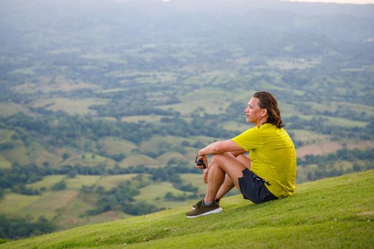 In summer, a man with long hair sits on the green slope of the mountain, looking into the distance at the valley. Dominican Republic, sunset in the mountains