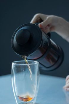 french press black tea pouring into a glass with a double bottom on a white table and grey background - vertical photo