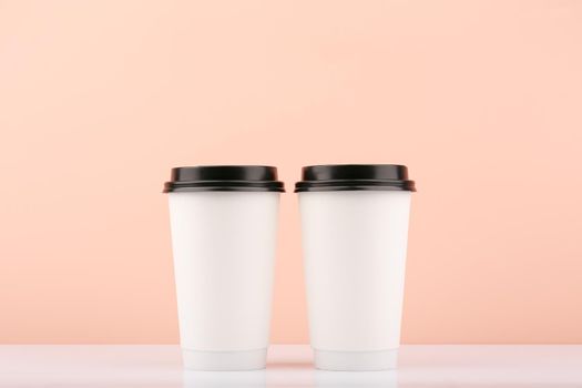 Two white disposable carton coffee or tea cups with black caps on white table against beige background with copy space. Concept of hot drinks