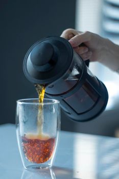 french press black tea pouring into a glass with a double bottom on a white table and grey background - vertical photo