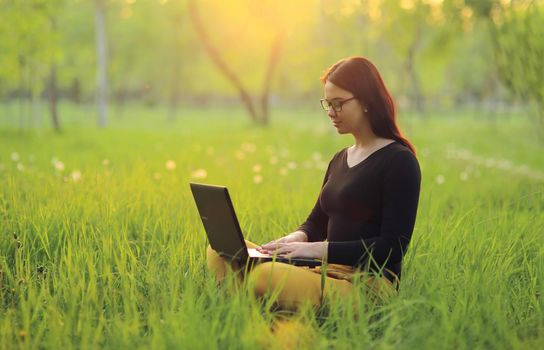 A cute smiling girl with a laptop sits on green grass in the park and works remotely as a freelancer. 