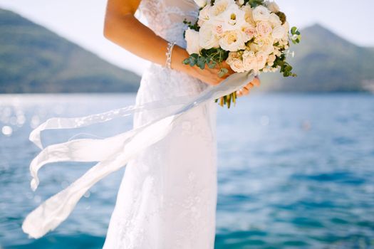 The bride in a white dress holds a beautiful wedding bouquet of roses in her hands on the background of the seascape. High quality photo