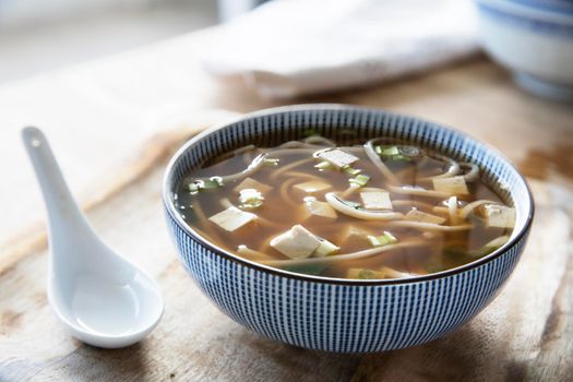 Bowl of red miso soup with tofu, green onions and udon noodles and white soup spoon.